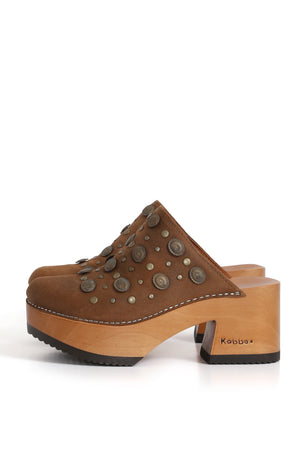 Sandalia tipo zueco Kebba Pájaro Pastora en cuero renobuk tostado, con detalle de tachas frontales. Planta de madera álamo de 8 cm de altura y forro en cuero de chancho caramelo. Fotografía sobre fondo blanco. Piezas únicas en colecciones limitadas. Hecho a mano en Villarrica. Kebba: tradición y herencia en tus manos.