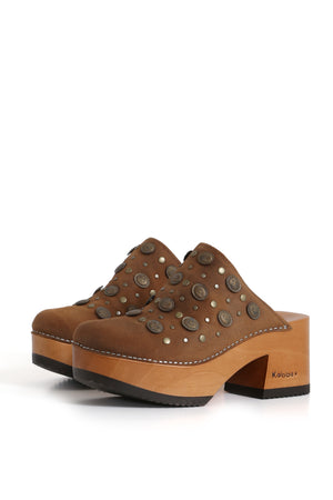 Kebba Pájaro Pastora, sandalia tipo zueco de cuero renobuk tostado con tachas metálicas en la parte frontal. Planta anatómica de madera álamo de 8 cm y forro de cuero de chancho caramelo. Imagen tres cuartos sobre fondo blanco. Piezas únicas en colecciones limitadas. Hecho a mano en Villarrica. Kebba: tradición y herencia en tus mano